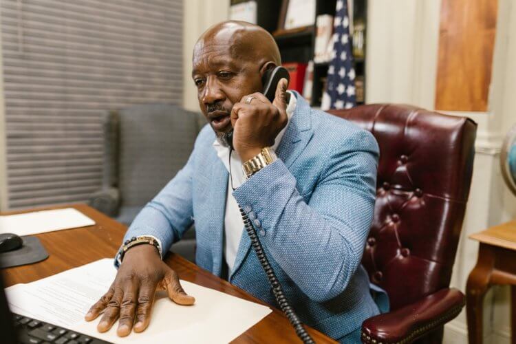 A male-presenting lawyer sitting at a desk on the phone with his legal malpractice carrier updating them about a legal malpractice claim